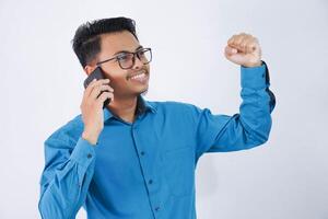retrato de emocionado o contento hermoso joven asiático empresario en vistiendo azul camisa demostración éxito gesto apretando puños mientras hablando en teléfono inteligente aislado en blanco antecedentes foto
