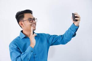 sonriente o contento asiático hombre con lentes participación teléfono inteligente para selfie foto vistiendo azul camisa aislado en blanco antecedentes