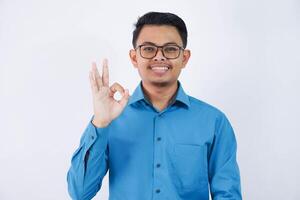 hermoso asiático masculino empleado con lentes sonriente en dedo gesto Okay o hecho vistiendo azul camisa aislado en blanco antecedentes foto