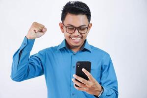 emocionado joven asiático empleado hombre con lentes participación teléfono con apretado puños vistiendo azul camisa aislado en blanco antecedentes foto