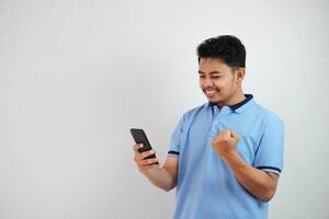 emocionado o contento retrato joven asiático hombre participación teléfono con apretado puños vistiendo azul polo t camisa aislado en blanco antecedentes foto