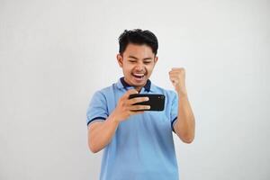 emocionado o contento joven asiático hombre ganar jugando juego con teléfono inteligente y con apretado puños aislado en blanco antecedentes foto