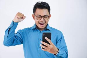 very excited young asian employee man with glasses holding phone with clenched fists wearing blue shirt isolated on white background photo