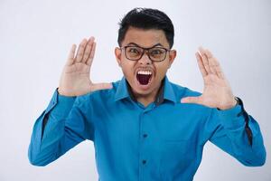 Annoyed handsome young asian man with glasses in wearing blue shirt raises palms while shouting loudly isolated on white background photo