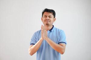 Young asian man wearing casual blue t shirt over white background begging with hands together with hope expression on face very emotional and worried. begging. sad face photo