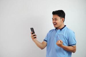 emocionado o contento retrato joven asiático hombre participación teléfono con apretado puños vistiendo azul polo t camisa aislado en blanco antecedentes foto