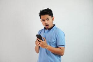 young Asian man with astonished expression looking dissatisfied with annoyed face while opening his mouth and holding phone wearing blue t shirt isolated on white background photo