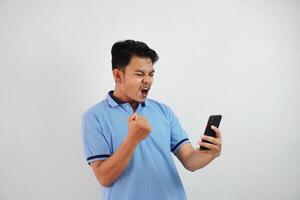 emocionado o contento retrato joven asiático hombre participación teléfono con apretado puños vistiendo azul polo t camisa aislado en blanco antecedentes foto