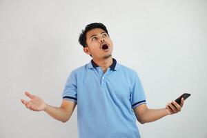 young Asian man with astonished expression looking dissatisfied with annoyed face while opening his mouth and holding phone wearing blue t shirt isolated on white background photo