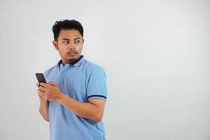 Surprised standing young asian man wearing blue t shirt with holding smartphone, trying to dodge isolated on white background photo