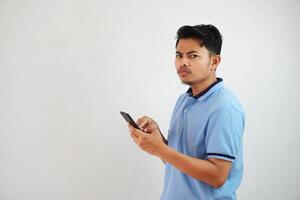 joven asiático hombre en pie participación un teléfono con un enojado y desaprobando expresión vistiendo azul t camisa aislado en blanco antecedentes foto