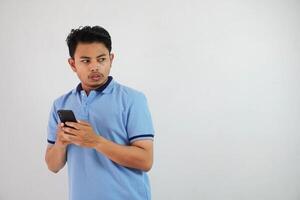 sorprendido en pie joven asiático hombre vistiendo azul t camisa con participación teléfono inteligente, molesto a esquivar aislado en blanco antecedentes foto