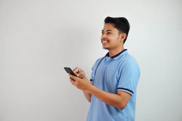 smiling or happy young asian man wearing blue t shirt holding phone and pointing at smartphone with finger, isolated on white background