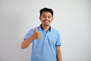 retrato de alegre asiático hombre en vistiendo azul t camisa sonriente y demostración pulgares arriba a cámara aislado terminado blanco antecedentes foto