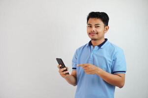 sonriente o contento joven asiático hombre vistiendo azul t camisa participación teléfono y señalando a teléfono inteligente con dedo, aislado en blanco antecedentes foto