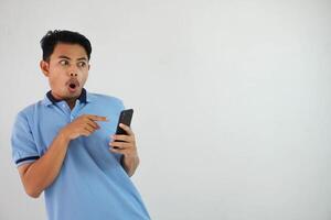 retrato asiático hombre conmocionado participación teléfono y señalando a el teléfono con un dedo vistiendo azul polo t camisa aislado en blanco antecedentes foto