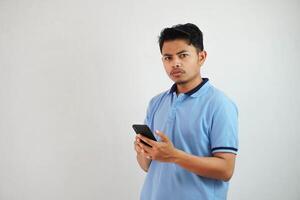 joven asiático hombre en pie participación un teléfono con un enojado y desaprobando expresión vistiendo azul t camisa aislado en blanco antecedentes foto