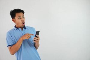 retrato asiático hombre conmocionado participación teléfono y señalando a el teléfono con un dedo vistiendo azul polo t camisa aislado en blanco antecedentes foto
