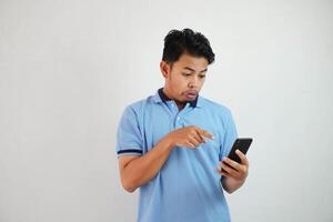 retrato asiático hombre conmocionado participación teléfono y señalando a el teléfono con un dedo vistiendo azul polo t camisa aislado en blanco antecedentes foto