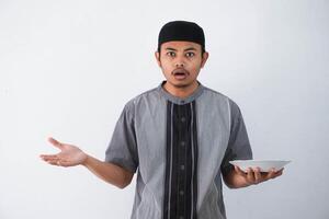 young asian muslim man showing disappointed expression while showing empty dinner plate wearing grey muslim clothes isolated on white background photo