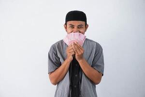 shocked young asian muslim man holding paper money gift THR from family ramadan Eid al-Fitr time wearing grey muslim clothes isolated on white background photo