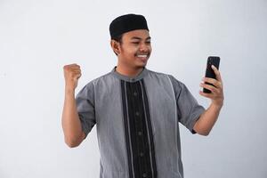 sonriente o contento joven asiático musulmán hombre participación teléfono inteligente con mano puño haciendo victorioso gesto, contento obtener especial regalo en línea, aislado en blanco antecedentes foto