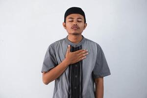 young Asian muslim man wearing grey muslim clothes with cap hands on chest with closed eyes and grateful gesture on face isolated on white background. health concept. photo