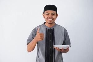 sonriente o contento joven asiático musulmán hombre demostración emocionado expresión dando pulgar arriba mientras participación vacío cena plato vistiendo gris musulmán ropa aislado en blanco antecedentes foto