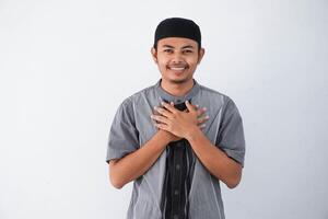 young Asian muslim man wearing grey muslim clothes with cap smiling with hands on chest with closed eyes and grateful gesture on face isolated on white background. health concept. photo