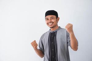 Successful happy young asian muslim man screaming shouting and shows winning victory gesture wearing grey muslim clothes isolated on white background photo