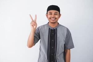 smiling or happy young asian muslim man showing pointing up with fingers number two while confident and showing peace sign with fingers wearing koko clothes isolated on white background photo