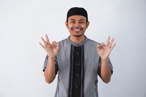 Young handsome asian muslim man smiling positive doing ok sign with hand and fingers wearing grey koko clothes isolated on white background. Successful expression. photo