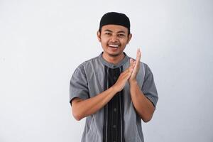 Smiling young Asian Muslim man clapping in hands and looking at camera wearing grey koko clothes isolated on white background photo