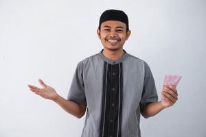 smiling or happy young asian muslim man holding and pointing paper money gift THR from family ramadan Eid al-Fitr time wearing grey muslim clothes isolated on white background photo