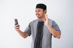 portrait of handsome young asian muslim man wearing koko clothes and using smartphone taking a video call and waving smile at phone isolated on white background photo