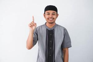 joven asiático musulmán hombre dedo señalando arriba levantamiento un dedo tiene un bueno idea mira sorprendido con un sonrisa vistiendo gris koko ropa, aislado en un blanco antecedentes. foto