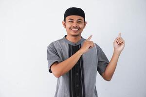 happy young asian muslim man pointing something on his side wearing grey muslim clothes isolated on white background photo