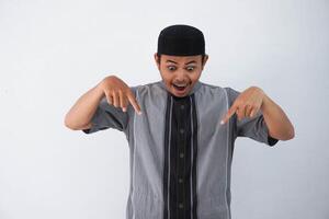 shocked young asian muslim man open mouth and amazed, looking and pointing downwards in disbelief and surprise wearing grey koko clothes isolated on white background photo