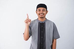 young asian muslim man finger pointing up raising a finger got a good idea looks surprised with a smile wearing grey koko clothes, isolated on a white background. photo