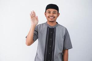 sonriente o contento joven asiático musulmán hombre abierto mano mientras ondulación mano decir Hola vistiendo gris musulmán ropa aislado en blanco antecedentes foto