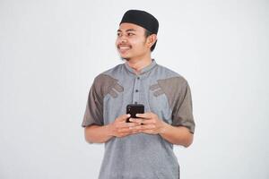 contento o sonriente joven asiático musulmán hombre participación teléfono celular vistiendo koko ropa aislado en blanco antecedentes. Ramadán concepto foto