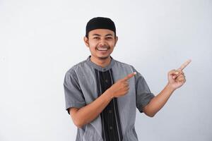 happy young asian muslim man pointing something on his side with his both hand wearing grey muslim clothes isolated on white background photo