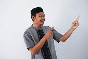 happy young asian muslim man pointing something on his side with his both hand wearing grey muslim clothes isolated on white background photo