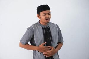 young asian muslim man squeeze his stomach for having stomachache pain wearing grey muslim clothes isolated on white background photo