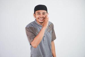 smiling young Asian Muslim man wearing grey muslim cloth, hand beside mouth whispering secrets or gossips isolated on white background photo