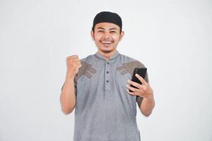 Excited or happy young Asian Muslim man holding on mobile phone and hands in fists doing winner gesture wearing koko clothes isolated over white background photo