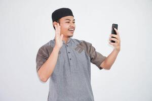 portrait of handsome young asian muslim man wearing koko clothes and using smartphone taking a video call and waving smile at phone isolated on white background photo