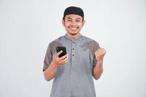 Excited or happy young Asian Muslim man holding on mobile phone and hands in fists doing winner gesture wearing koko clothes isolated over white background photo