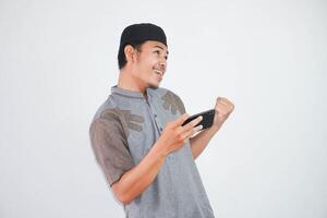 Excited or happy young Asian Muslim man holding on mobile phone and hands in fists doing winner gesture wearing koko clothes isolated over white background photo