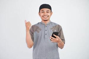 smiling or happy asian muslim man holding phone with thumbs up wearing grey muslim clothes isolated on white background photo